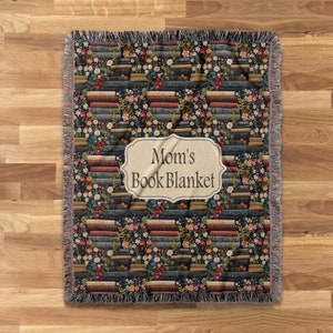 May include: A woven throw blanket with a pattern of stacked books and flowers. The blanket has a label that reads "Mom's Book Blanket".