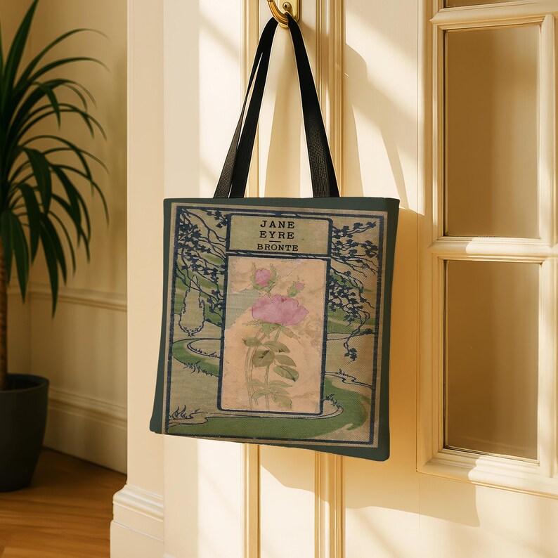 May include: A rectangular tote bag with black shoulder straps featuring a printed book cover design. The graphic displays the text JANE EYRE BRONTE above a central illustration of a pink rose on a beige background, framed by green foliage.