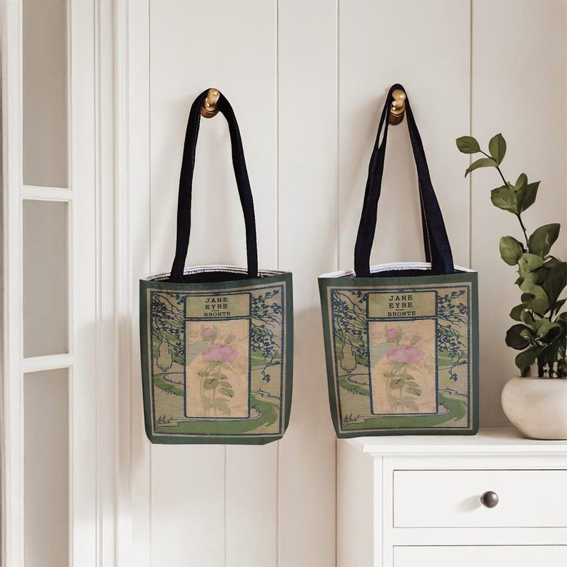 May include: Two identical tote bags with black handles, featuring a rectangular graphic print of a pink rose illustration, green foliage, and the text JANE EYRE BRONTE centered on a light tan background with a dark green border.