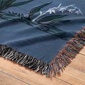 May include: A blue woven throw blanket with a floral pattern and fringe trim. The blanket is folded and lying on a wooden surface.