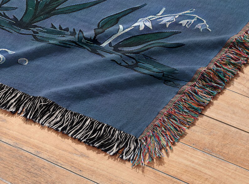 May include: A blue woven throw blanket with a floral pattern and fringe trim. The blanket is folded and lying on a wooden surface.