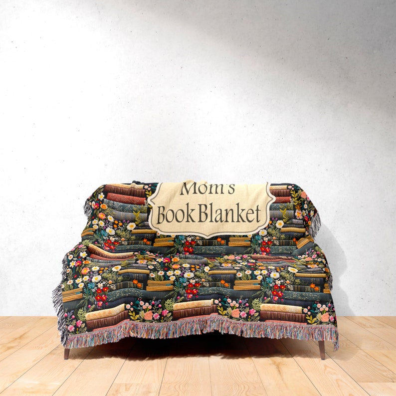 May include: A woven throw blanket with a pattern of stacked books and flowers. The blanket has a cream-colored label with the text "Mom's Book Blanket".