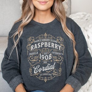 May include: A dark gray sweatshirt with a vintage-style graphic design featuring the text "Prince Edward Island Raspberry" and "Cordial" with the words "Marilla", "Est. 1908", and "Cuthbert's" in smaller text.