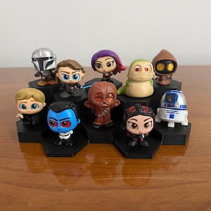 May include: A collection of Star Wars character figurines, including The Mandalorian, Anakin Skywalker, and R2-D2, displayed on a black hexagonal tiered stand. The figures are brightly colored and detailed, showcasing various characters from the franchise.