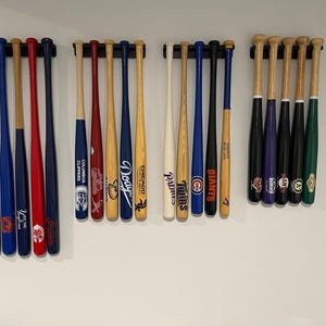 May include: A collection of baseball bats in various colors, including blue, red, black, and natural wood tones. Some bats feature team logos and text. The bats are mounted on a white wall.