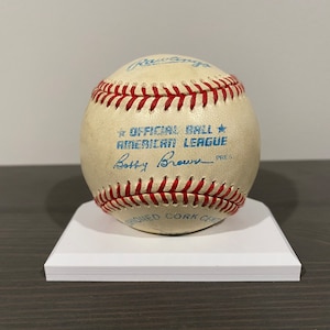 May include: A white baseball with red stitching and blue text that reads "OFFICIAL BALL AMERICAN LEAGUE" and "Bobby Brown - PRES".
