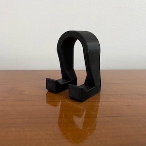 May include: A black, U-shaped phone stand. The stand has two rectangular feet and a curved top, designed to hold a phone upright. The stand is made of a solid, matte black material and sits on a wooden surface.