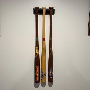 May include: Three baseball bats displayed on a wall mount. The bats are brown and natural wood, with colorful graphics and text. The bats are hanging vertically, showcasing their length and design.