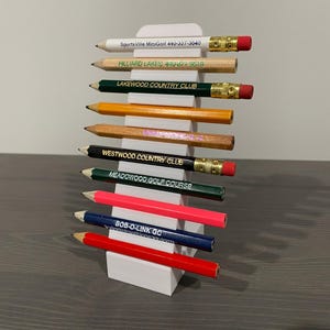May include: A collection of pencils with various golf course and business names printed on them, including "SportsVille MiniGolf" and "Lakewood Country Club." The pencils are displayed in a white holder.