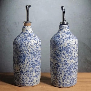 May include: Two ceramic oil dispensers featuring a speckled blue and white pattern. Each bottle has a cylindrical body, a narrow neck, and a metallic pour spout. One dispenser includes a cork stopper, while the other has a black cap.