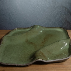 May include: A square ceramic tray with an irregular, wavy rim and a glossy sage green glaze. The surface features subtle brown speckles and a textured, dark brown edge. The tray has a slightly undulating base with two distinct recessed sections.