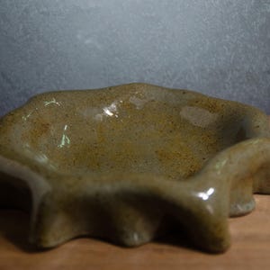 May include: A shallow, hand-formed ceramic bowl with a glossy, speckled olive-green glaze. The rim features a distinct, undulating, wavy edge that creates a series of rounded protrusions around the entire circumference of the circular vessel.