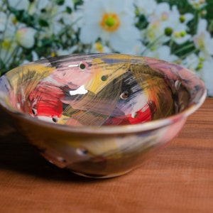 May include: A ceramic bowl with a colourful abstract design. The bowl features a mix of red, yellow, black, and pink brushstrokes. It has several small holes around the rim. The bowl is set on a wooden surface.