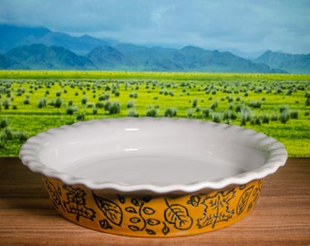 May include: A ceramic pie dish with a white interior and a scalloped edge. The exterior is yellow with black leaf and vine designs. The dish sits on a wooden surface with a blurred background of a green field and mountains.