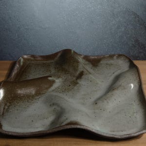 May include: A square ceramic tray with an irregular, wavy surface. The piece features a mottled glaze with a blend of dark brown, earthy tones and a lighter, speckled grey-white centre, creating a textured appearance with raised ridges.