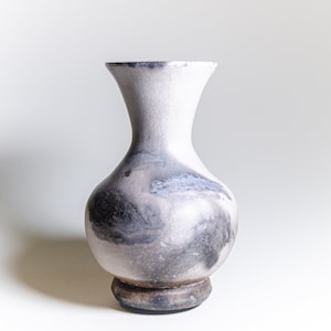 May include: A ceramic vase with a bulbous body and a flared neck. The vase has a light gray and white color scheme with dark gray and black accents. It sits on a small, round, dark brown base.