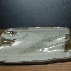 May include: A rectangular ceramic tray with an uneven, wavy surface featuring a glossy off-white glaze with dark brown speckles. The edges of the tray are dark brown and textured, contrasting with the smooth, undulating center of the piece.