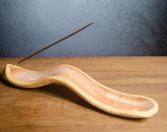 May include: A handmade ceramic incense holder with a wavy design. The holder is a light yellow color with a pinkish-orange interior. An incense stick is placed in the holder, which sits on a wooden surface.