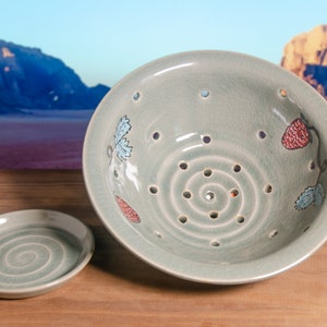 May include: A pale green ceramic colander and small dish set. The colander features a spiral pattern of holes and is decorated with painted strawberries and leaves. The small dish has a matching spiral design.
