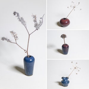 May include: A collage of four small ceramic vases in shades of blue and burgundy, each holding a dried plant stem. The vases vary in shape, including a round, a cylindrical, and a fluted design. The background is plain white.