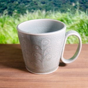 May include: A light gray ceramic mug with a handle, featuring a crackle glaze and a decorative swirl pattern. The mug sits on a wooden surface, with a blurred green background. A simple, elegant design for beverages.