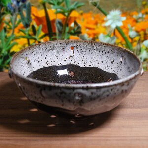 May include: A handmade ceramic bowl with a speckled white and dark brown glaze. The bowl has a rounded shape with small holes near the rim. The interior is a glossy dark brown. The bowl is sitting on a wooden surface.