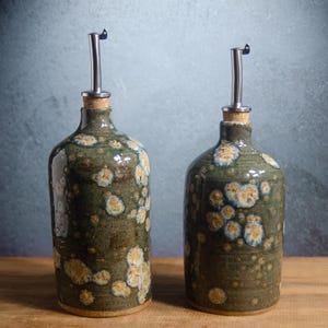 May include: Two cylindrical ceramic oil dispensers with a dark olive green glaze featuring light cream and tan crystalline patterns. Each bottle is topped with a cork stopper and a silver-colored stainless steel pour spout for liquid dispensing.