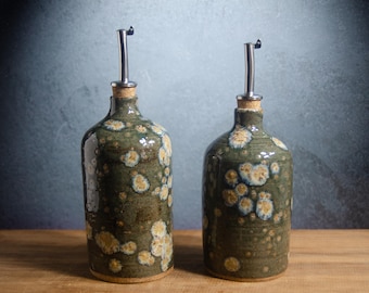 May include: Two cylindrical ceramic oil dispensers with a dark olive green glaze featuring light cream and tan crystalline patterns. Each bottle is topped with a cork stopper and a silver-colored metal pour spout with a small black cap.