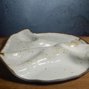 May include: A ceramic serving platter with an off-white, speckled glaze and a dark brown, uneven rim. The platter features a distinct, undulating, wave-like surface with three separate, shallow, curved sections divided by raised ridges.