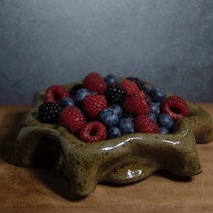 May include: A low-profile, star-shaped ceramic bowl with a speckled, olive-green glaze filled with a fresh assortment of red raspberries, dark blackberries, and small blue blueberries. The bowl features a thick, uneven rim and a glossy finish.
