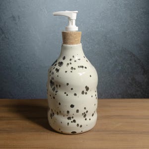 May include: A ceramic soap dispenser with a cream-colored glaze and black speckled pattern. The bottle has a rounded, slightly irregular shape and is topped with a natural cork stopper and a white plastic pump mechanism for dispensing liquid soap.