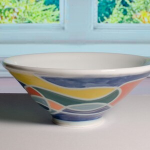 May include: A white ceramic bowl with a colorful abstract design. The bowl features a wide rim and a footed base. The exterior is decorated with blue, yellow, green, and pink curved shapes. The bowl is likely used for serving food.