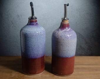 May include: Two ceramic oil dispensers with a cylindrical body, featuring a deep red base that transitions into a speckled light blue and purple glaze. Each bottle is topped with a metallic pour spout and a cork stopper for controlled dispensing.