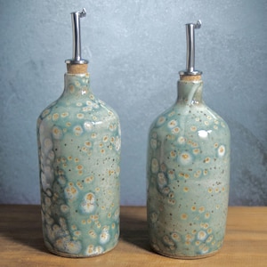 May include: Two ceramic oil bottles featuring a mottled teal and light blue glaze with white and tan circular spots. Each bottle has a narrow neck, a cork stopper, and a silver-colored metal pour spout with a small hinged lid at the top.