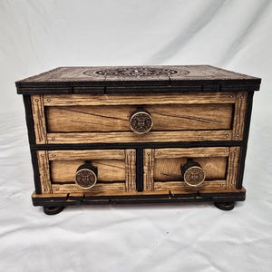 May include: A wooden chest with two drawers, featuring a sun-like design on the drawer pulls and top. The chest has a dark brown frame and light brown drawer fronts. The top is intricately carved with a circular pattern.