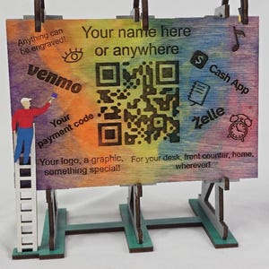 May include: A wooden sign with a rainbow-colored background and a large QR code. The sign includes the text "Your name here or anywhere," and logos for Venmo, Cash App, and Zelle. The sign is supported by a green base and has brown arched handles.