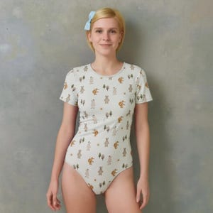 May include: A light green short-sleeve bodysuit featuring an all-over print of small brown bears, squirrels, and trees. The garment has a round neckline and a fitted silhouette, showcasing a repeating pattern of woodland animals and flora.