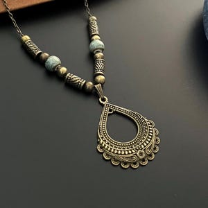 May include: A bronze necklace featuring a teardrop-shaped pendant. The pendant has an intricate design and is surrounded by smaller beads. The necklace is made of metal and has a rustic look.