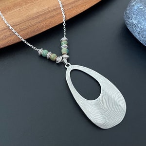 May include: A silver necklace with a large oval pendant featuring a textured, brushed metal design. The pendant has a cutout oval in the center. The necklace is adorned with green and brown agate beads.