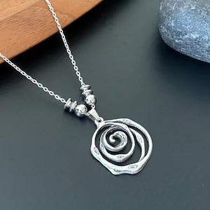 May include: A silver-toned necklace with a pendant featuring a swirling, openwork design. The pendant hangs from a delicate chain, accented with small, round beads. The necklace rests on a dark surface.