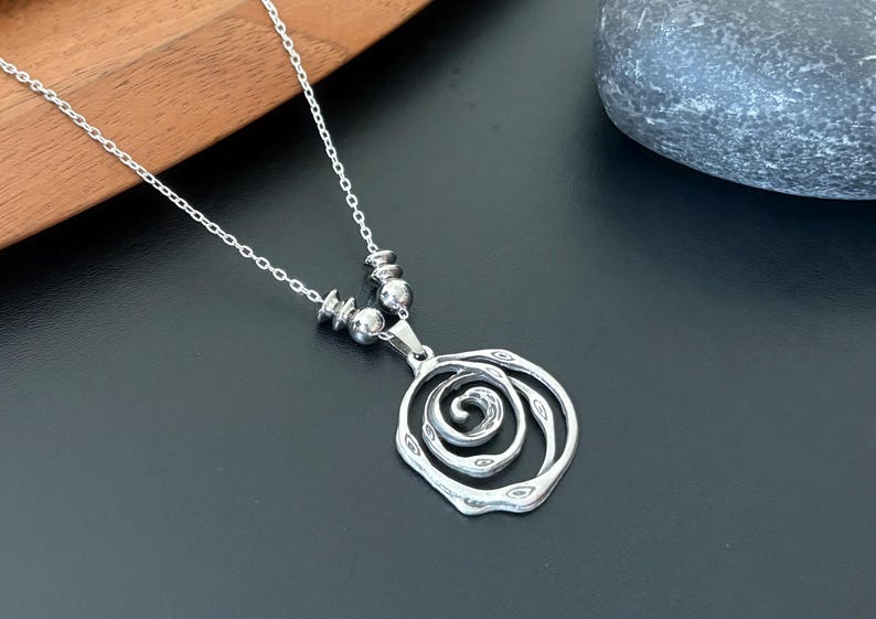 May include: A silver-toned necklace with a pendant featuring a swirling, openwork design. The pendant hangs from a delicate chain, accented with small, round beads. The necklace rests on a dark surface.
