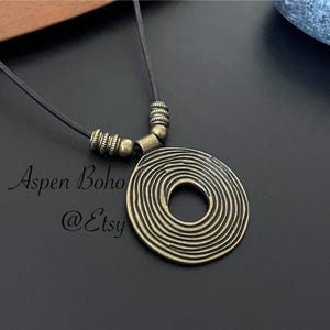 May include: A black leather cord necklace with a large, round, bronze pendant. The pendant has a concentric circle design.