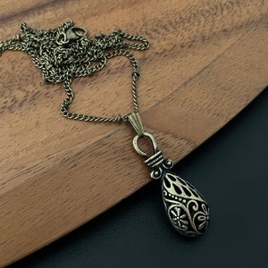 Könnte beinhalten: Eine goldfarbene Halskette mit einem detaillierten Tropfen-Anhänger. Der Anhänger hat eine schwarze Emaille-Oberfläche und komplizierte florale Designs.