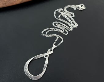 May include: A silver necklace with a textured, twisted pendant in the shape of a teardrop. The chain is a delicate, silver chain.