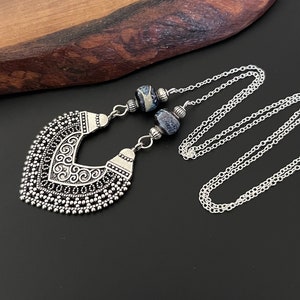 May include: A silver necklace featuring a large, ornate pendant and two blue and white agate beads. The pendant is heart-shaped with a decorative design.