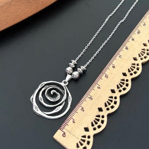May include: A silver-toned necklace with a spiral rose pendant. The pendant is connected to a chain with small, round beads. A wooden ruler is next to the necklace, showing measurements in inches.