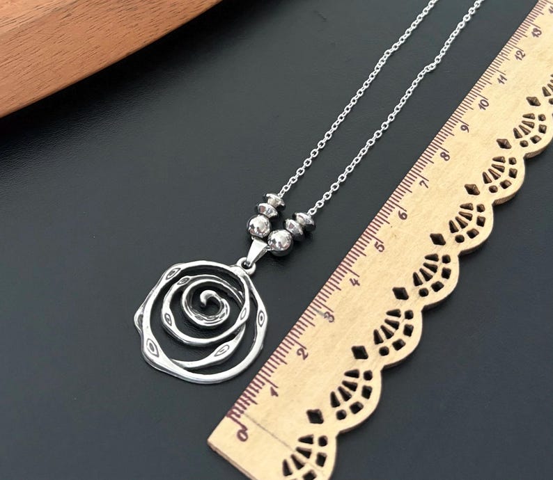 May include: A silver-toned necklace with a spiral rose pendant. The pendant is connected to a chain with small, round beads. A wooden ruler is next to the necklace, showing measurements in inches.