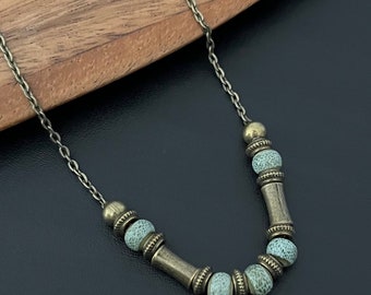 May include: A necklace with a bronze chain and decorative beads. The necklace features light blue, textured beads and bronze-colored cylindrical and round accents. The necklace is displayed on a wooden surface with a black background.