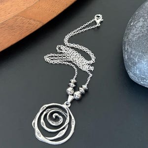 May include: A silver-toned necklace with a spiral pendant. The pendant has a layered, open design with a central spiral. The necklace chain is delicate, and the pendant is suspended from a series of small, round beads.