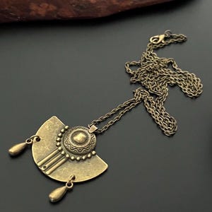 Könnte beinhalten: Eine goldfarbene Halskette mit einem großen, verzierten Anhänger. Der Anhänger hat die Form eines Halbkreises mit einem runden Design in der Mitte. Die Halskette hat einen Kettengliederstil.